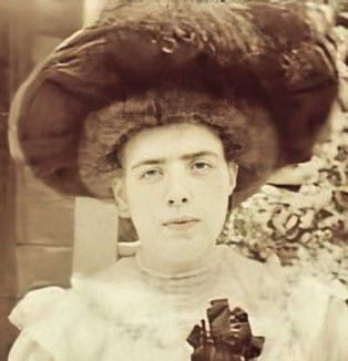 A headshot photo of a woman in a ruffled dress and huge feathered hat holding a flower.