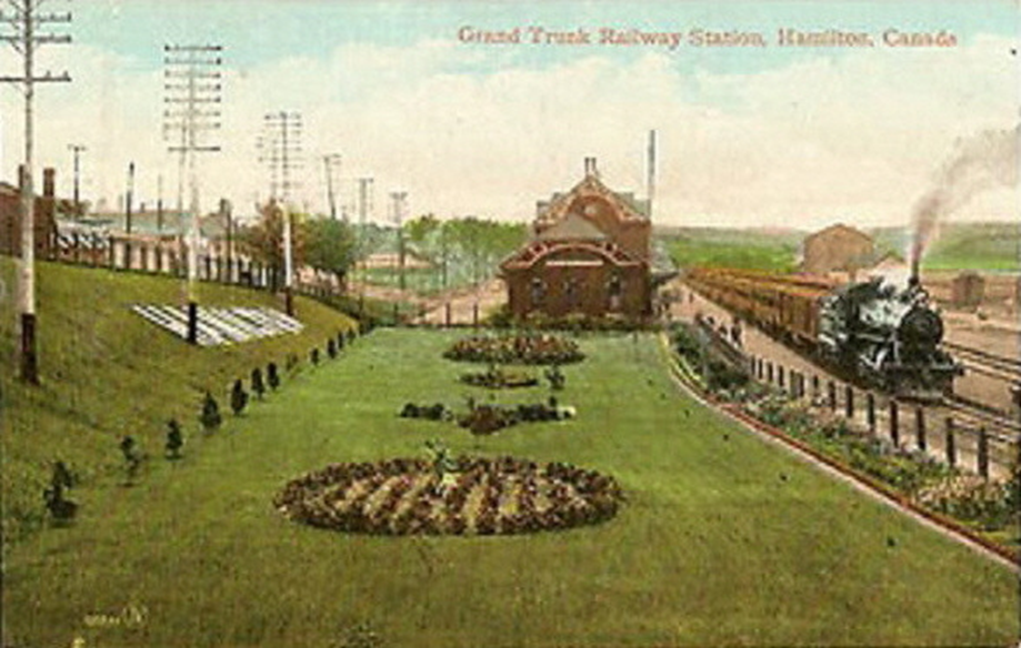 A colourized photo of the GTR Railway station in Hamilton (ca. 1903) with gardens and a train steaming past.