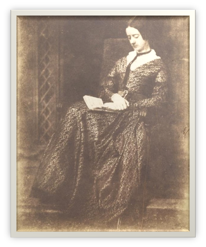 An early 19th century photo of a woman reading a book whilst seated in a floor-length dress.