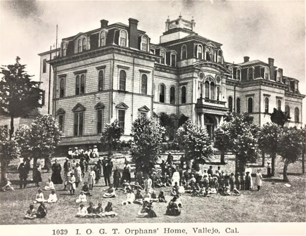 A photo of an institutional residence, with children and staff gathered beneath trees on the lawn.