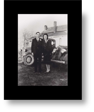 A man and woman pose together in front of 1940s vehicles parked at a house.