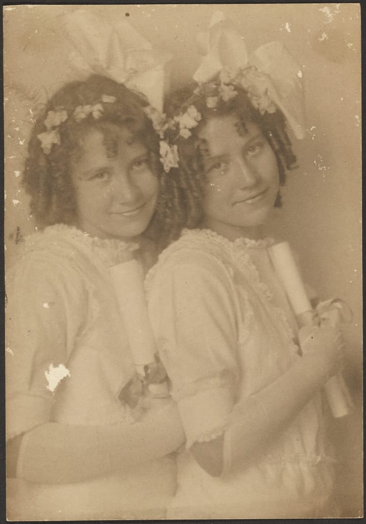 A old sepia-toned photograph of twin sisters, with ribbon and flower adorned curly hair, dressed in white.